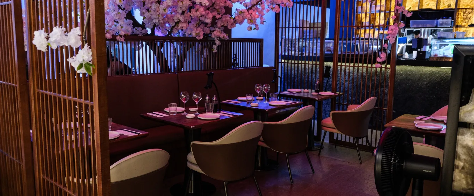 Intérieur élégant d'un restaurant avec des banquettes en cuir marron et des tables en bois sombre, décoré de cerisiers en fleurs artificiels roses et de cloisons en bois. On aperçoit un bar en arrière-plan et un ventilateur noir au premier plan à droite.