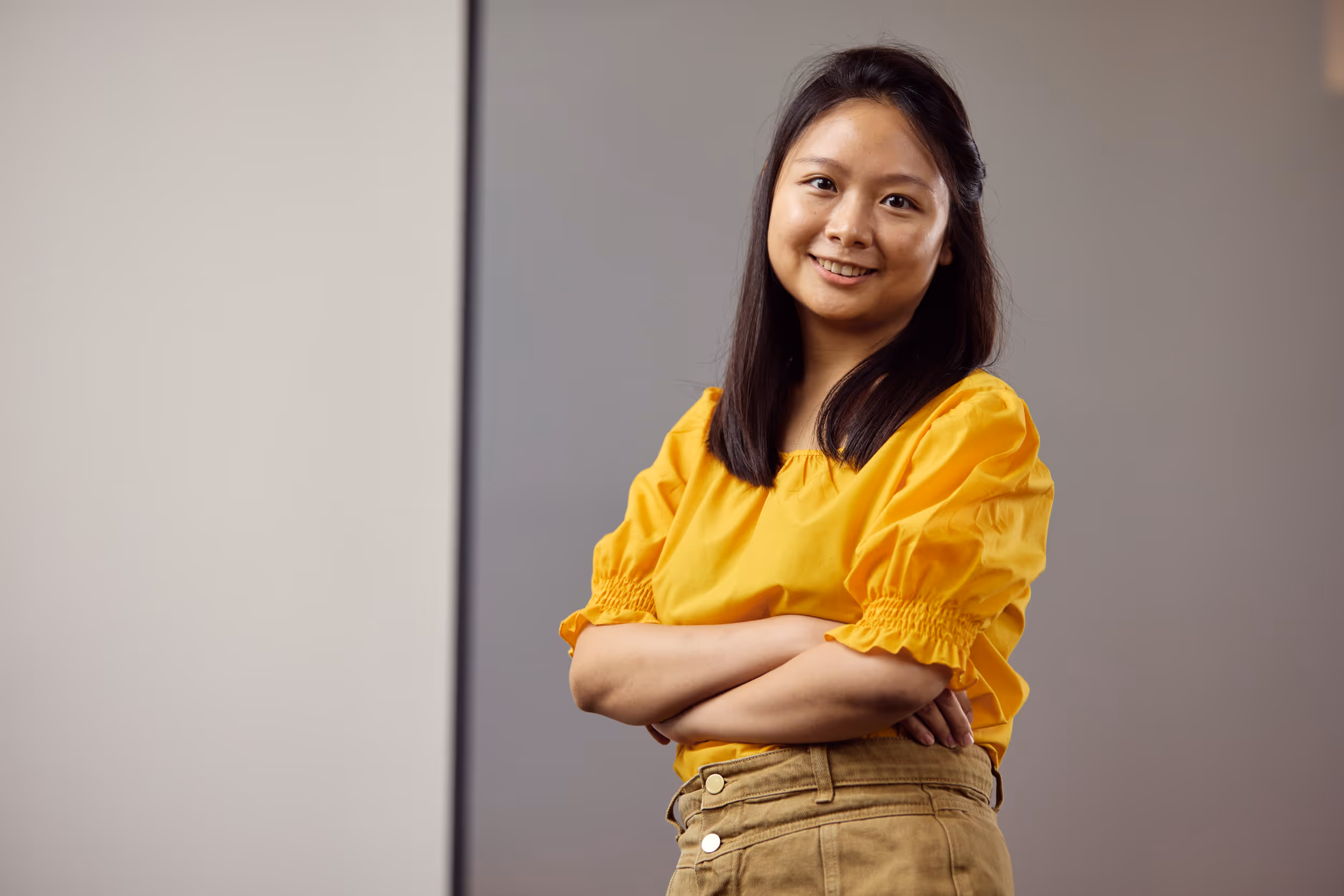 Young woman with long dark hair in a yellow blouse and tan pants standing with arms crossed, smiling against a gray background.