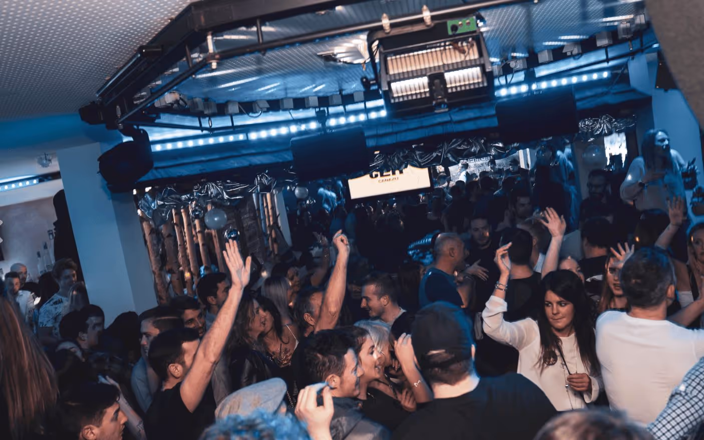 Crowded nightclub with people dancing and raising their hands under blue lighting.