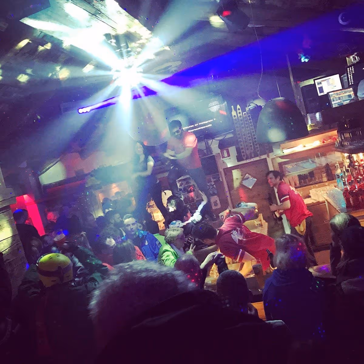 Crowded nightclub scene with colorful lights, people dancing and socializing under a bright disco light.
