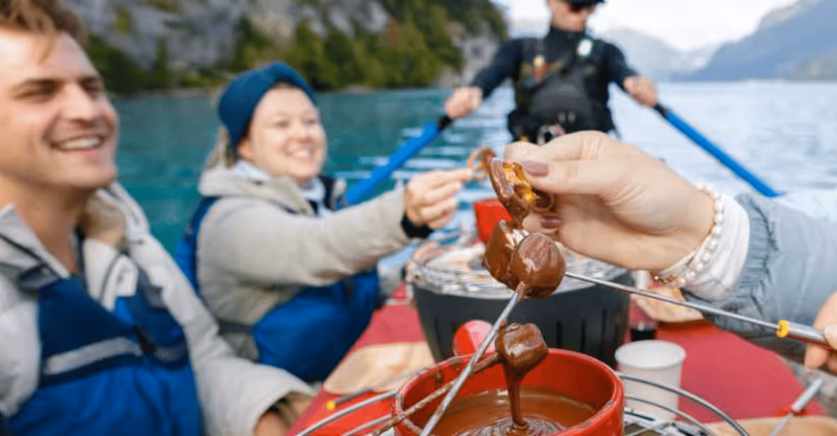 Group of people in life jackets enjoying dipping marshmallows into chocolate fondue on a boat with scenic mountain and lake background.