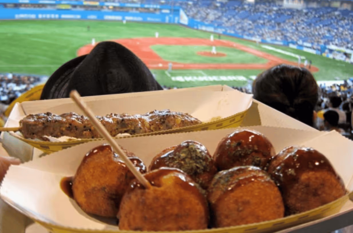Tray of skewered grilled meat and takoyaki balls with sauce, held up in a crowded baseball stadium.