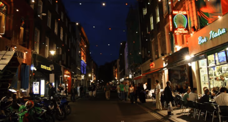Nighttime street scene with people walking and dining outdoors at lit restaurants and bars.