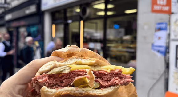 Hand holding a sandwich filled with sliced pastrami, pickles, and mustard in an outdoor urban setting.
