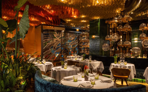 Elegant restaurant interior with round tables set with white tablecloths, green plants, and warm lighting from hanging glass fixtures.