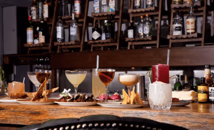 Assorted cocktails and appetizers arranged on a wooden bar counter with bottles and glasses displayed on shelves in the background.