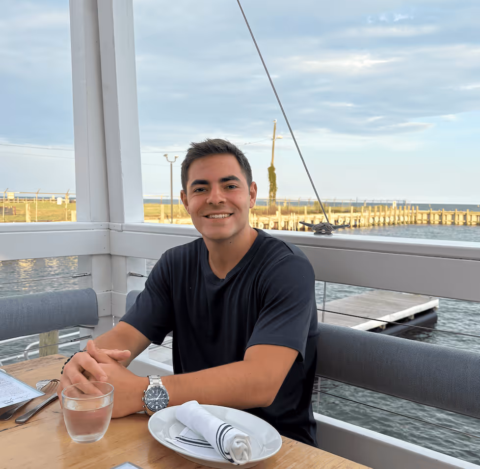Smiling young man in a black t-shirt sitting at a table with a glass of water and a plate with a rolled napkin, with a waterfront view in the background.