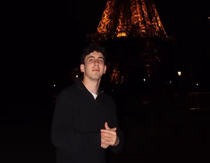 Young man in a black jacket posing at night with the illuminated Eiffel Tower in the background.