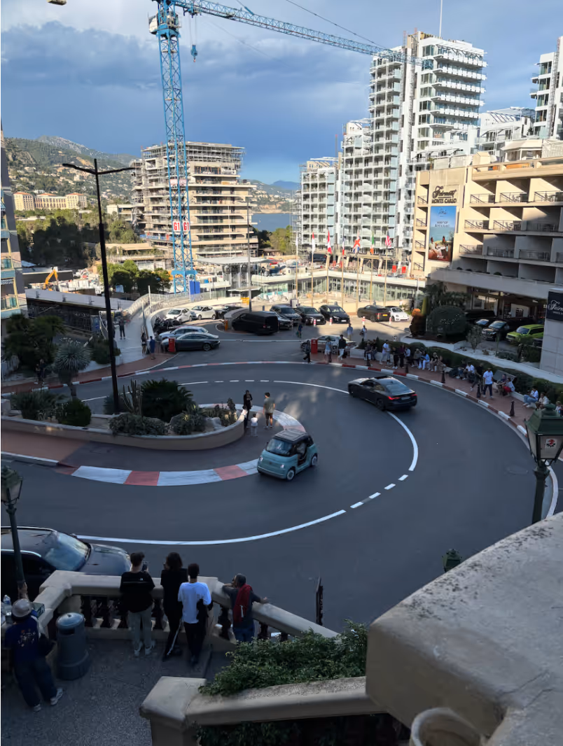 Curved street in an urban area with people watching cars navigate the sharp turn, surrounded by tall buildings and a construction crane.