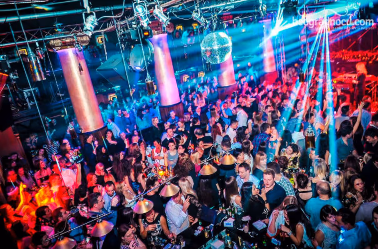 Crowded nightclub scene with people dancing and socializing under colorful lights and disco balls.
