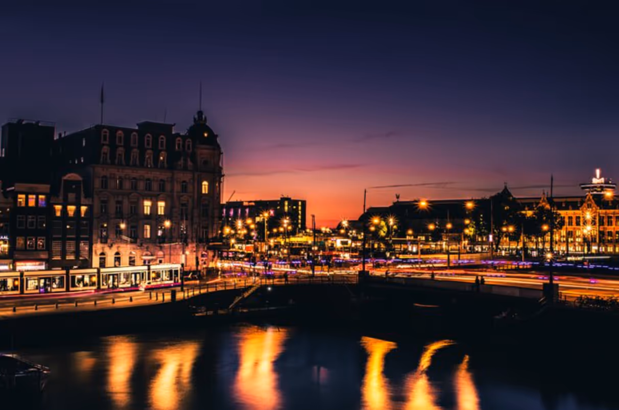 Evening cityscape with a river reflecting lights, historic buildings, and a vibrant sunset sky.