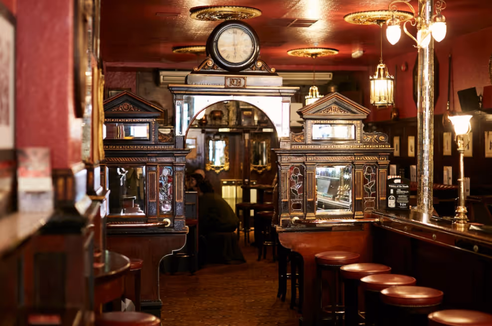 Interior of a vintage pub with wooden stools, ornate partition windows, and a large clock above an archway.
