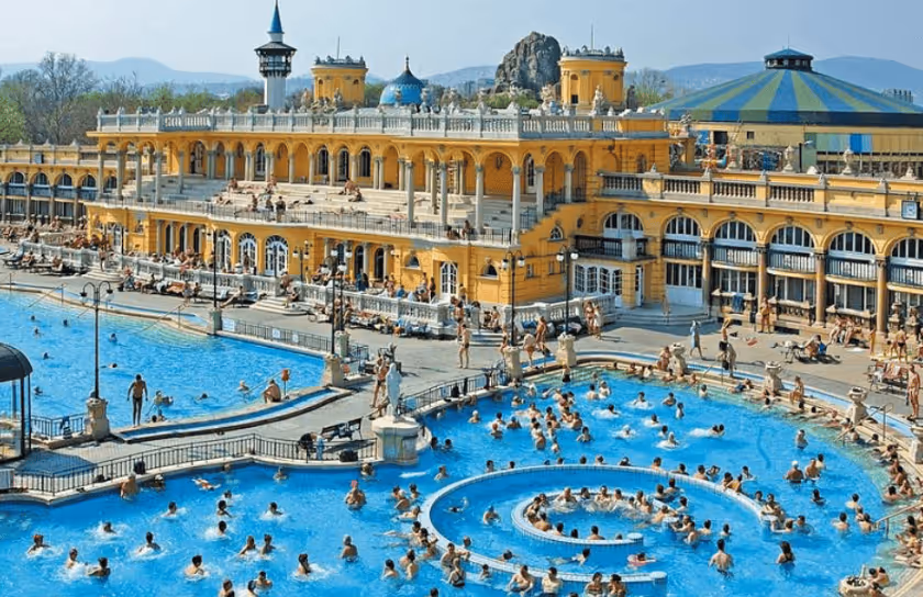 Large outdoor thermal baths with many people swimming and lounging, against a grand historic yellow building with arched windows and decorative towers.