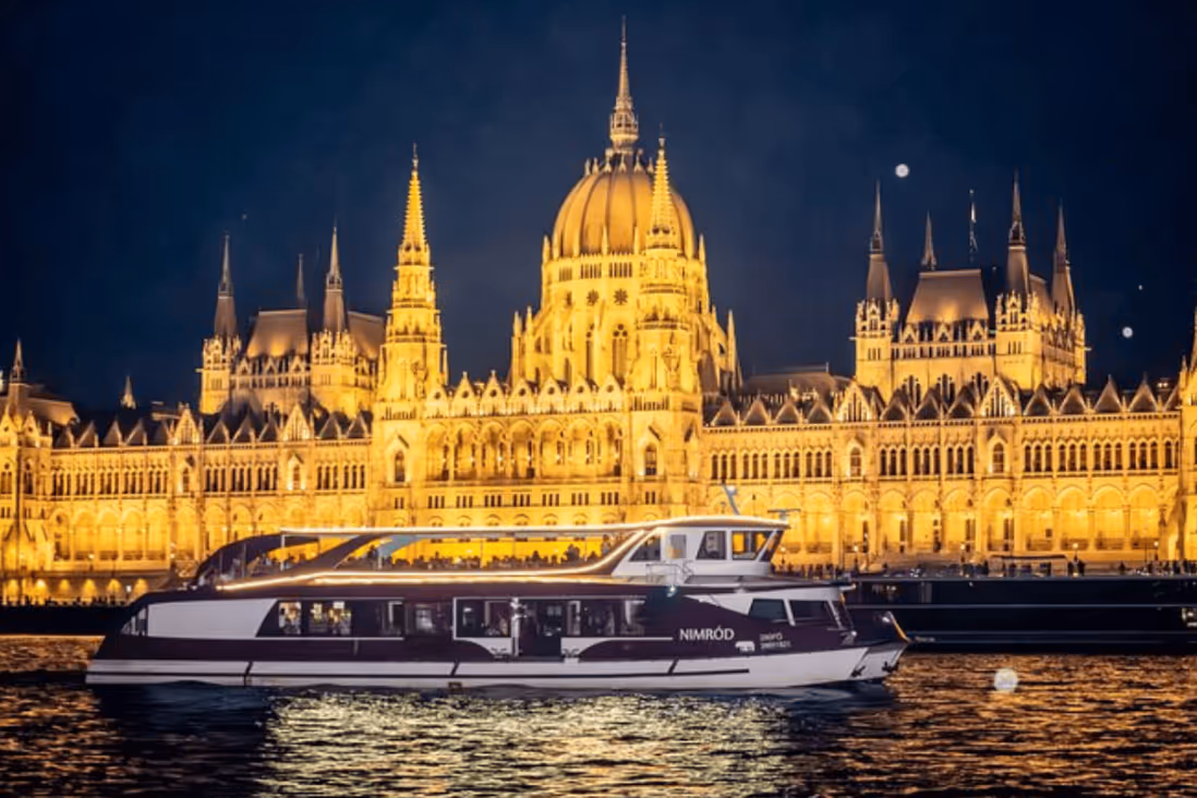 Night view of a modern passenger boat on the Danube River with the illuminated Hungarian Parliament building in the background.