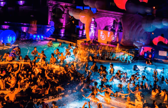 Crowded indoor pool party with many people swimming and socializing under vibrant purple and blue lighting.