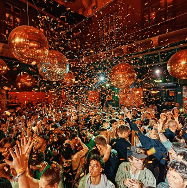 Crowded indoor party with people dancing and celebrating under large golden disco balls and confetti falling from above.