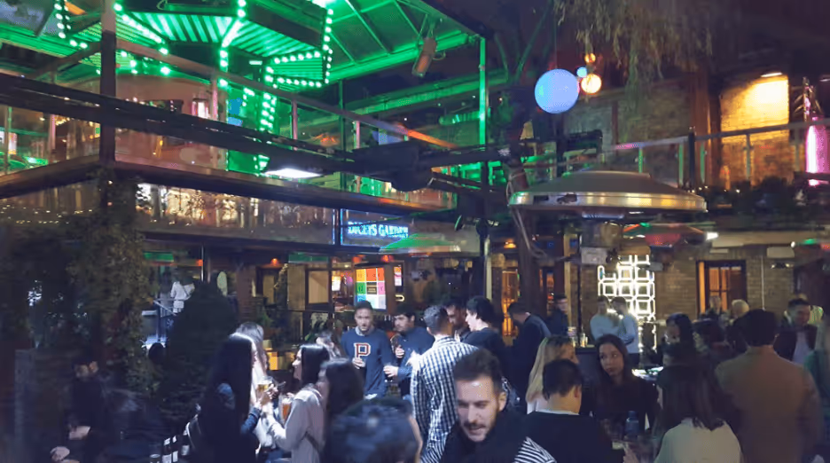 Crowded indoor bar with people socializing under green neon lights and industrial-style decor.