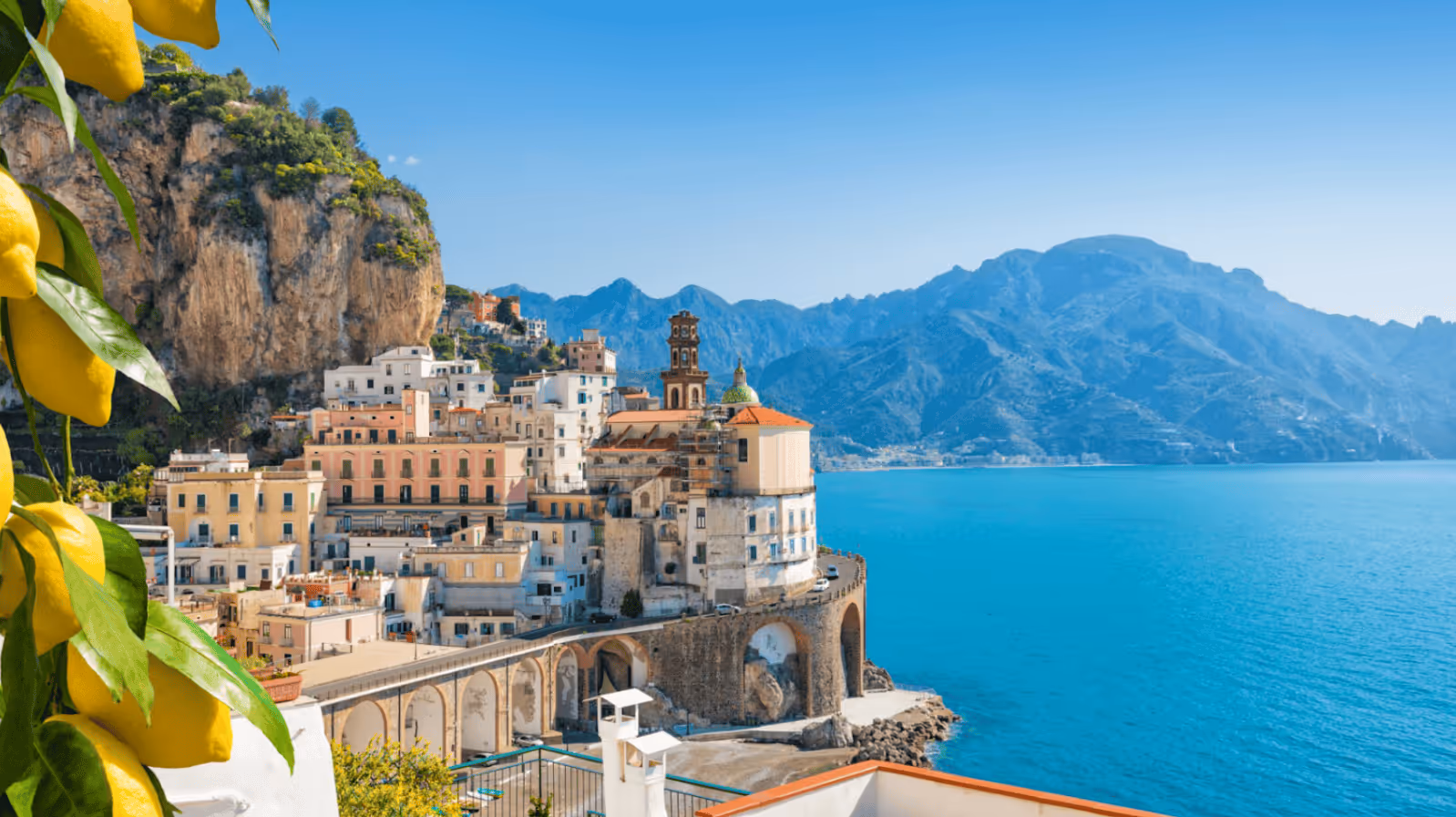 Coastal town with colorful buildings on cliffs beside bright blue sea and distant mountains under clear sky, with lemons in foreground.