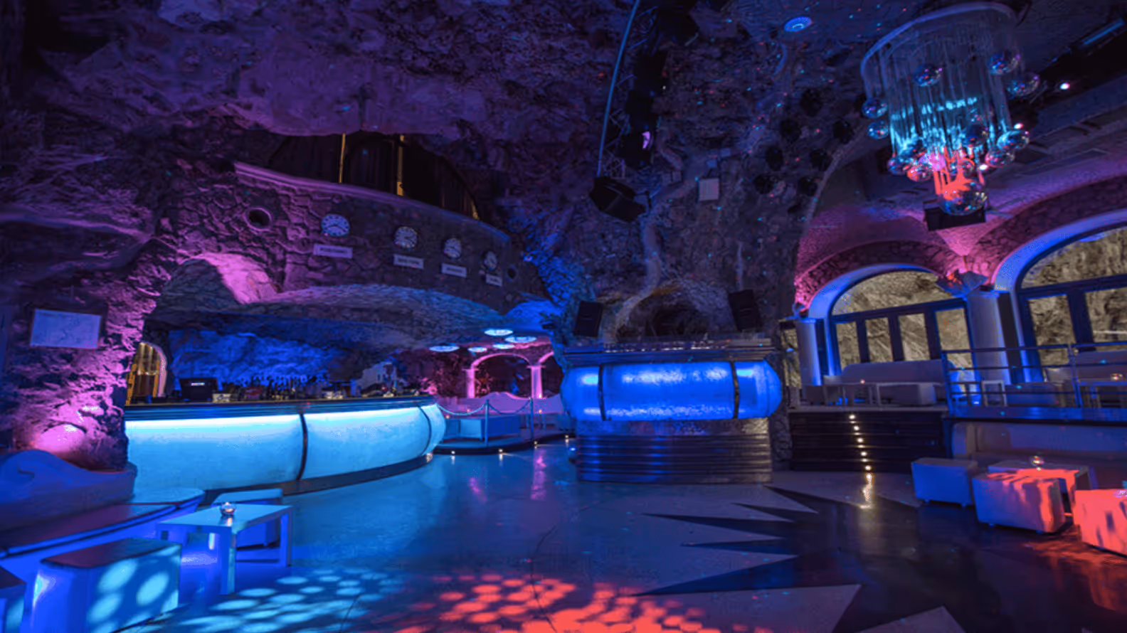 Nightclub interior with cave-like stone walls, illuminated bar counters, colorful purple and blue ambient lighting, and modern seating areas.