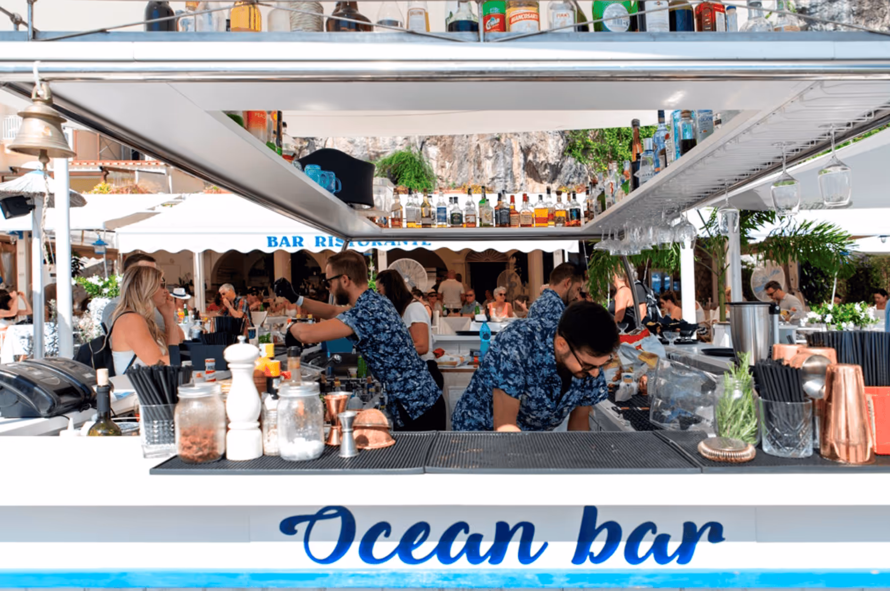 Bartenders in blue floral shirts preparing drinks at a busy outdoor Ocean Bar with customers and a rocky cliff in the background.