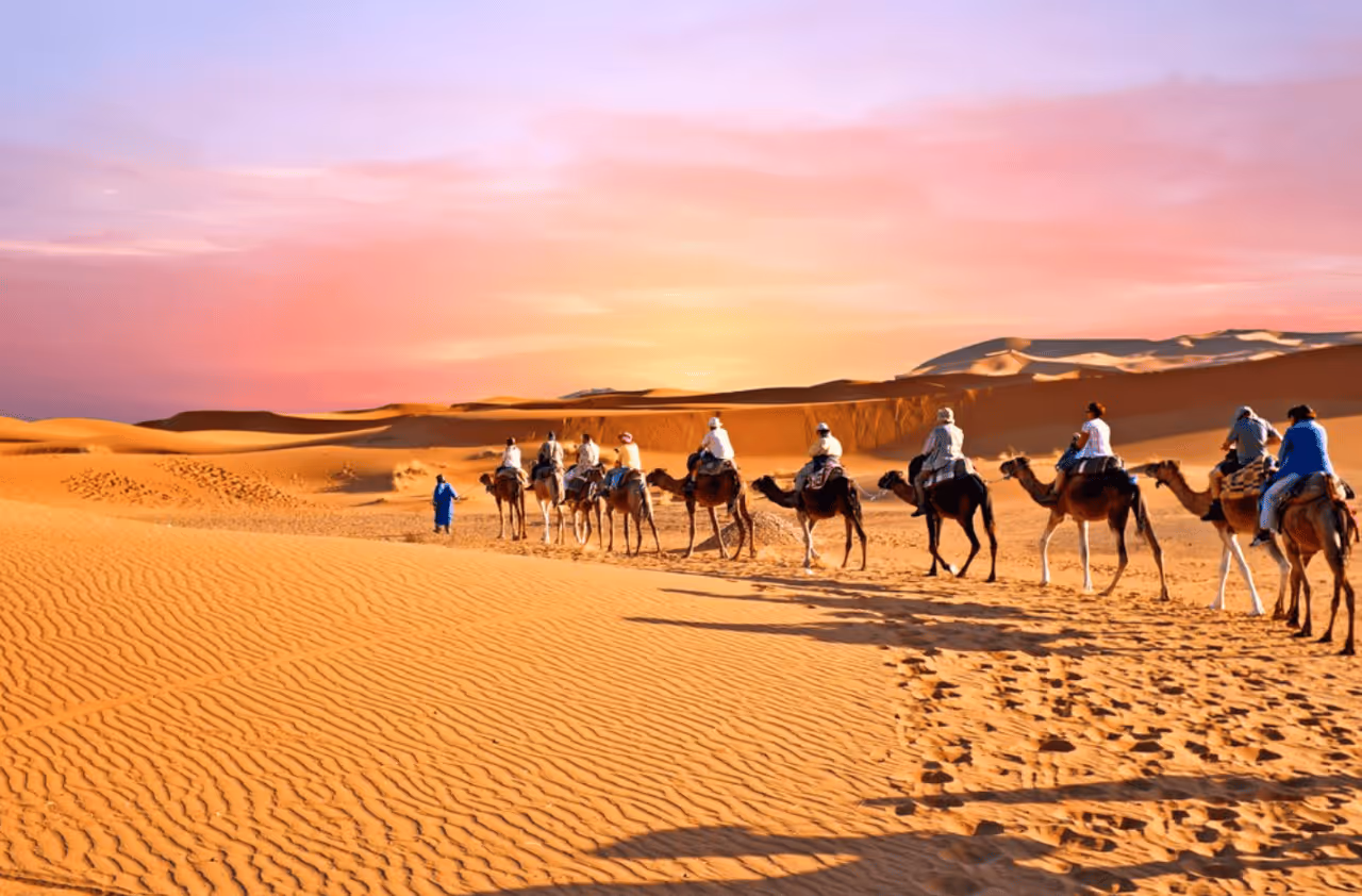 Group of people riding camels led by a person in blue walking through golden sand dunes at sunset.