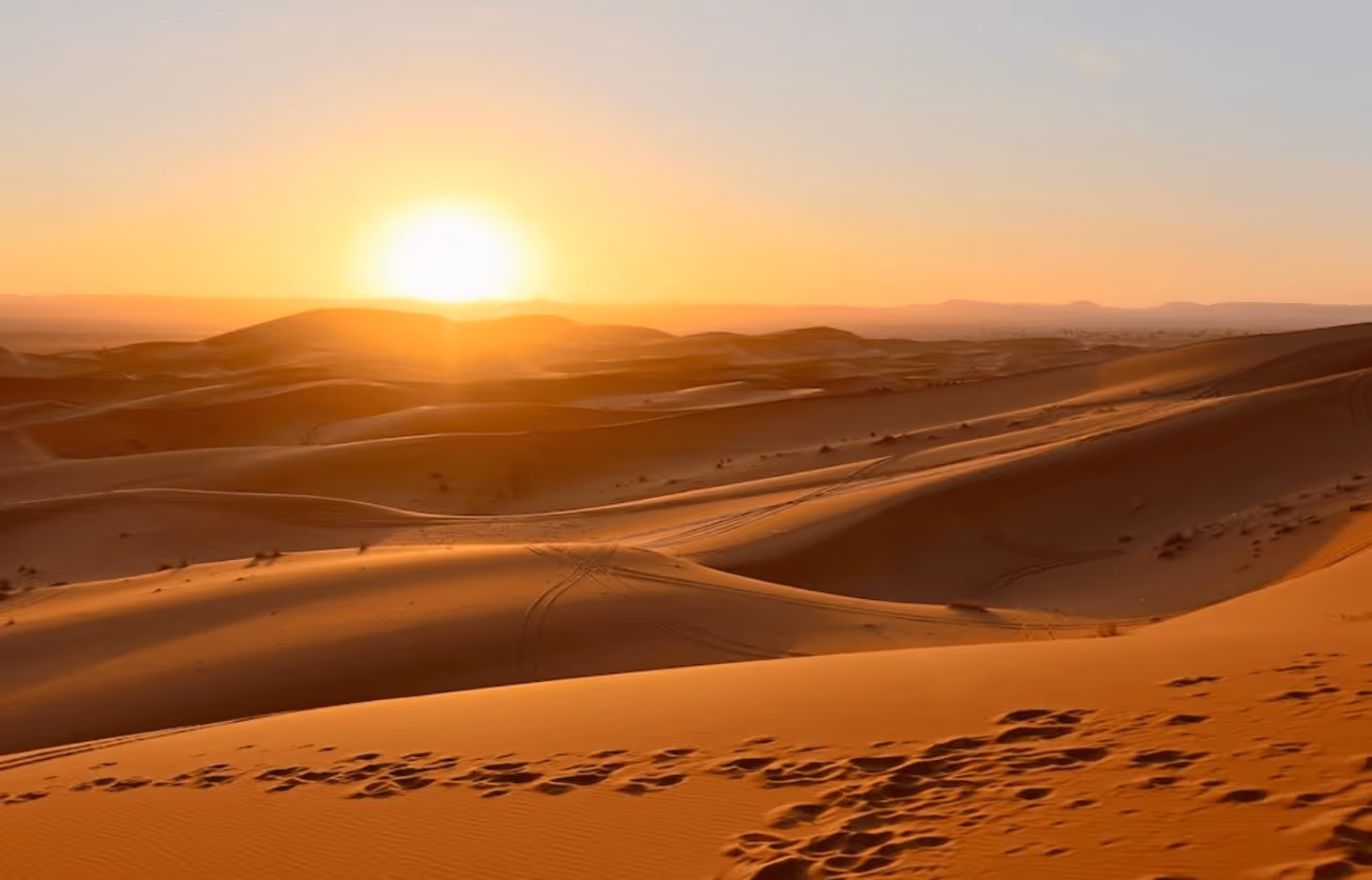 Sun setting over expansive sand dunes in a desert with footprints in the foreground.