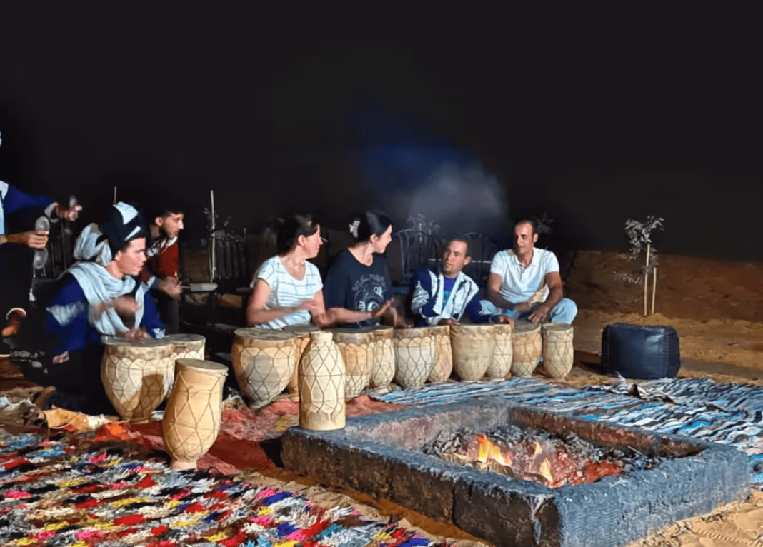 Group of people sitting around a fire pit at night playing traditional drums on colorful rugs in an outdoor desert setting.