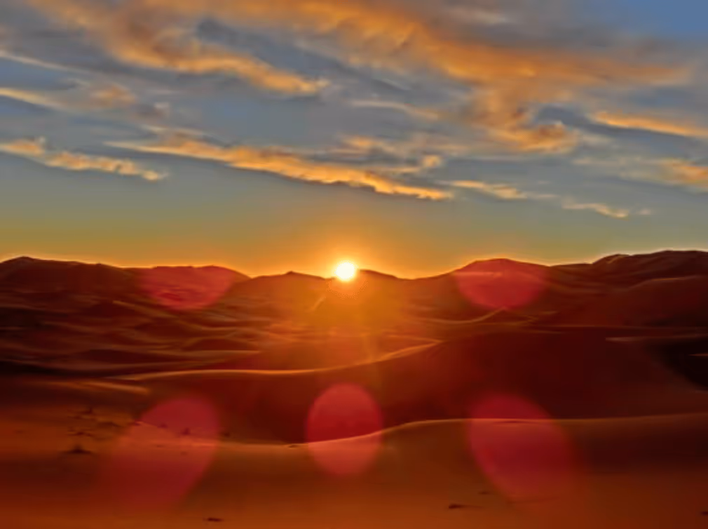 Sun setting behind mountain range with sandy dunes in the foreground and a partly cloudy sky.
