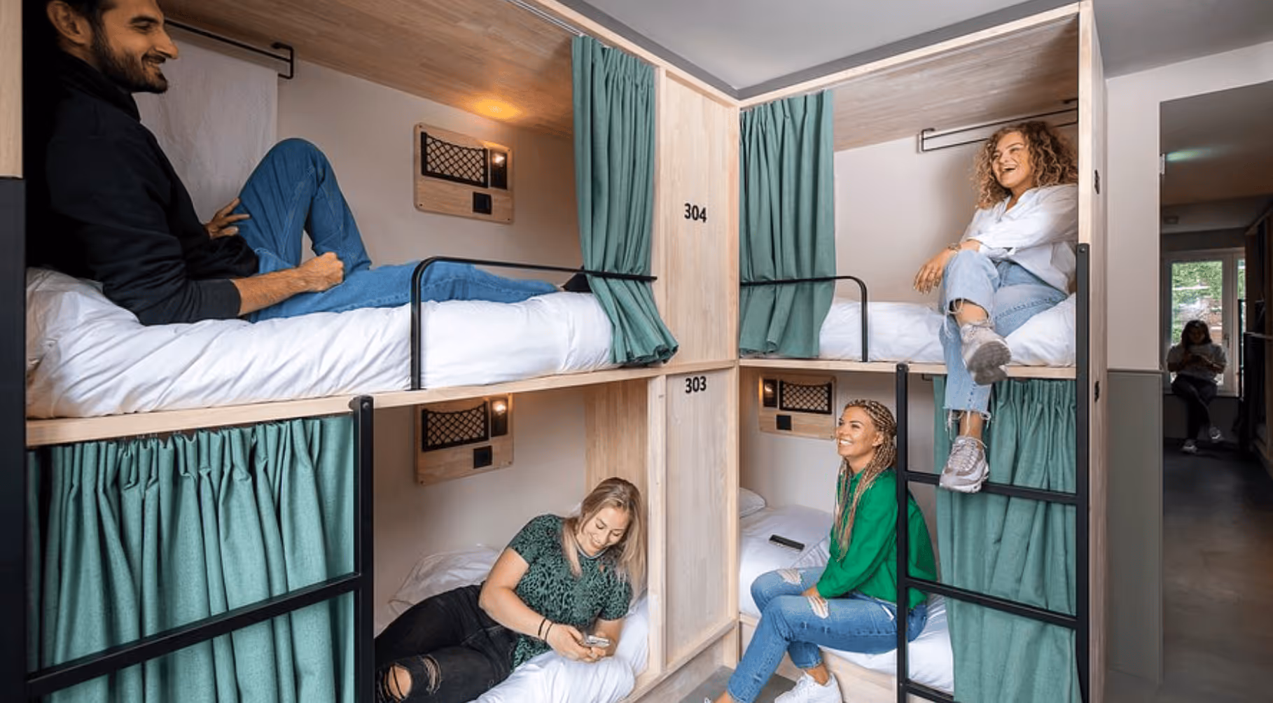 Four young adults relaxing and interacting in a modern capsule hostel room with stacked beds and green privacy curtains.