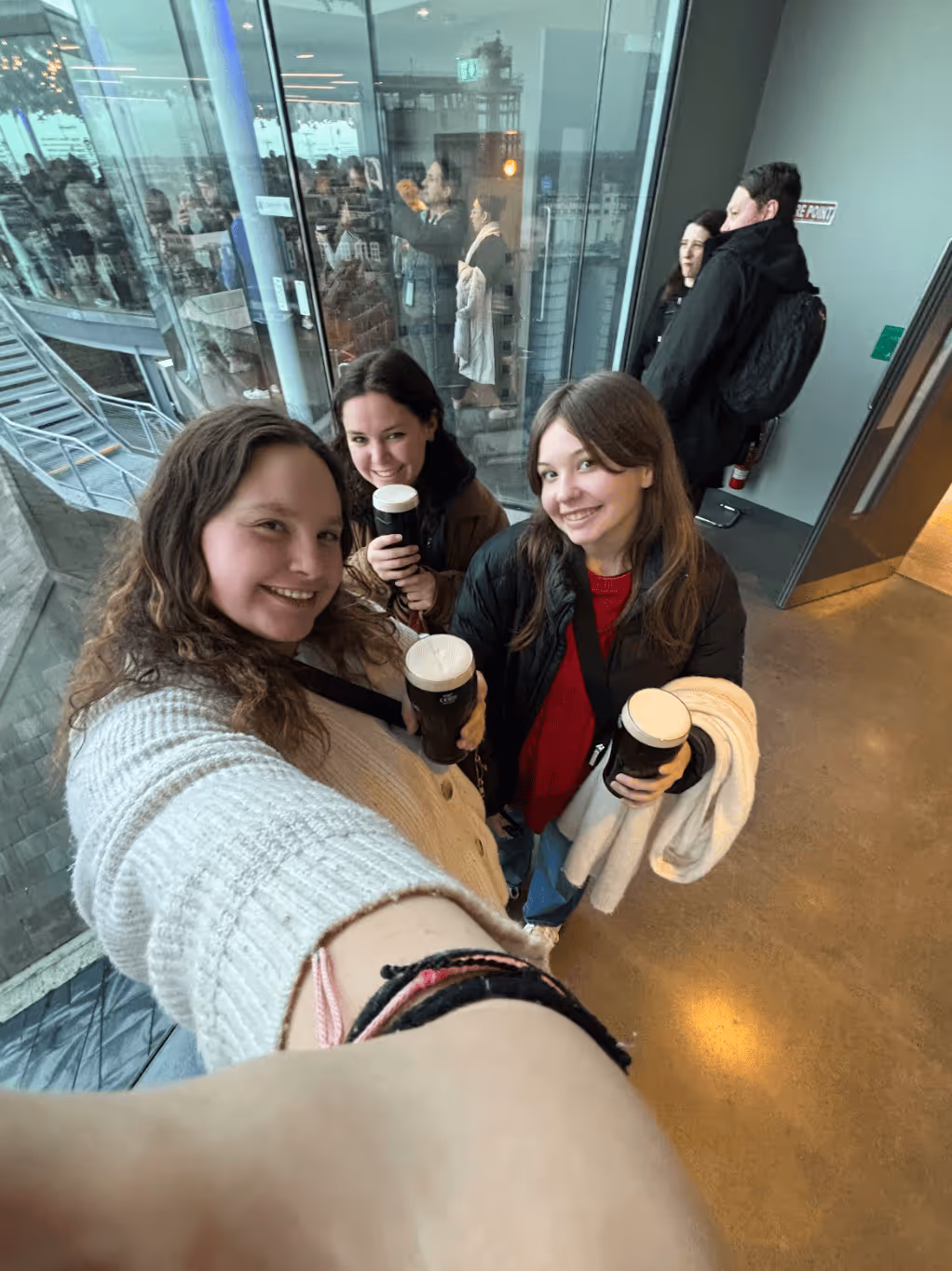 Three smiling women indoors holding pints of dark beer, taking a selfie near large windows.