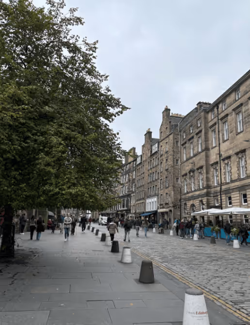 People walking along a tree-lined street with historic stone buildings and outdoor cafe seating on a cloudy day.