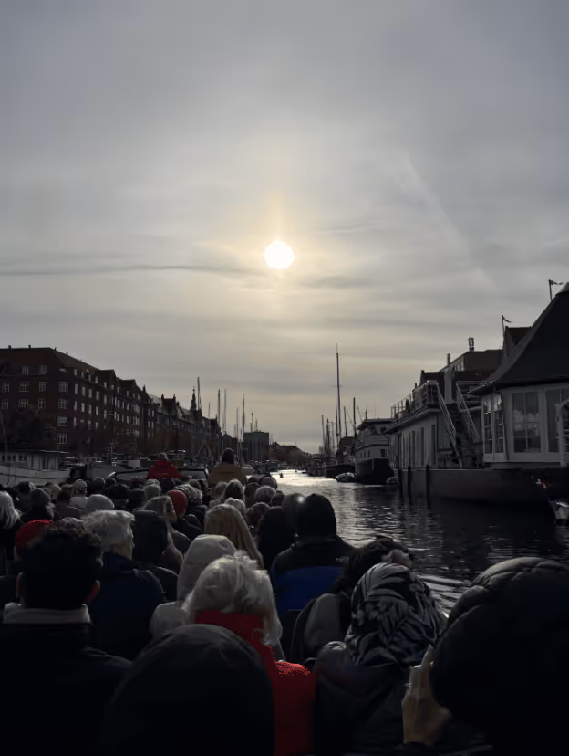 Crowded boat ride on a canal during sunset under a cloudy sky with buildings on both sides.