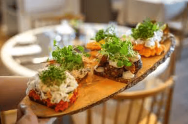 A wooden serving board with various gourmet appetizers topped with fresh greens, held by a person in a restaurant setting.