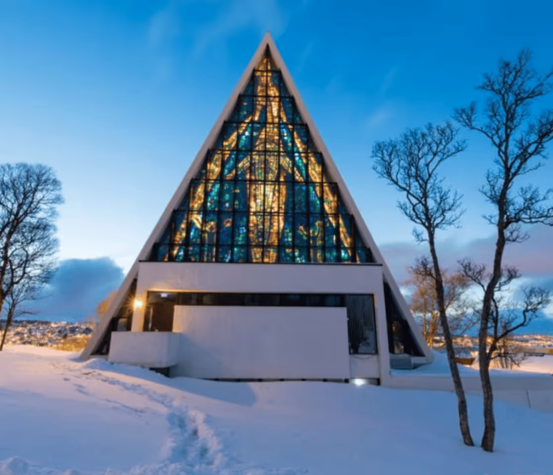 Modern triangular church with illuminated stained glass window surrounded by snow-covered ground and bare trees at dusk.