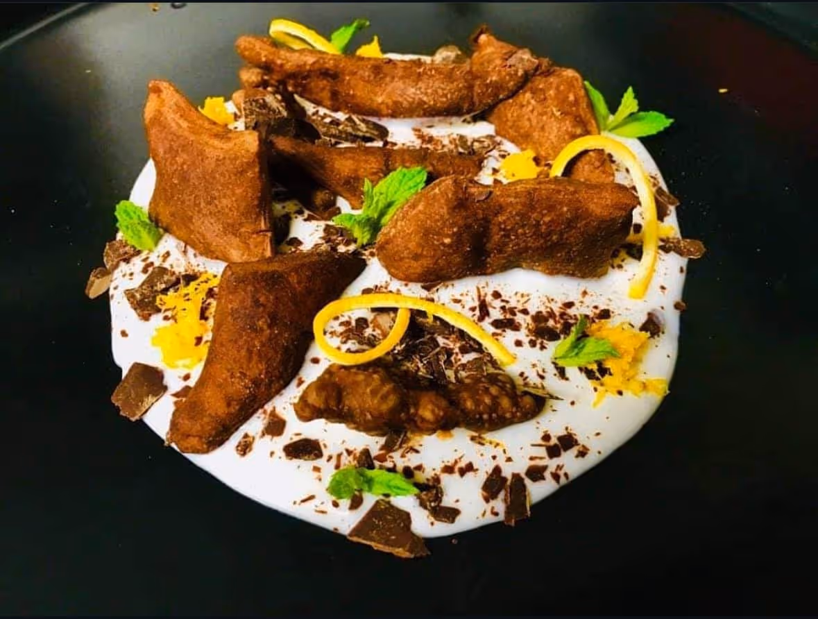 Chocolate-dusted fried pastries arranged on a white cream with chocolate shavings, lemon zest, and mint leaves on a black plate.