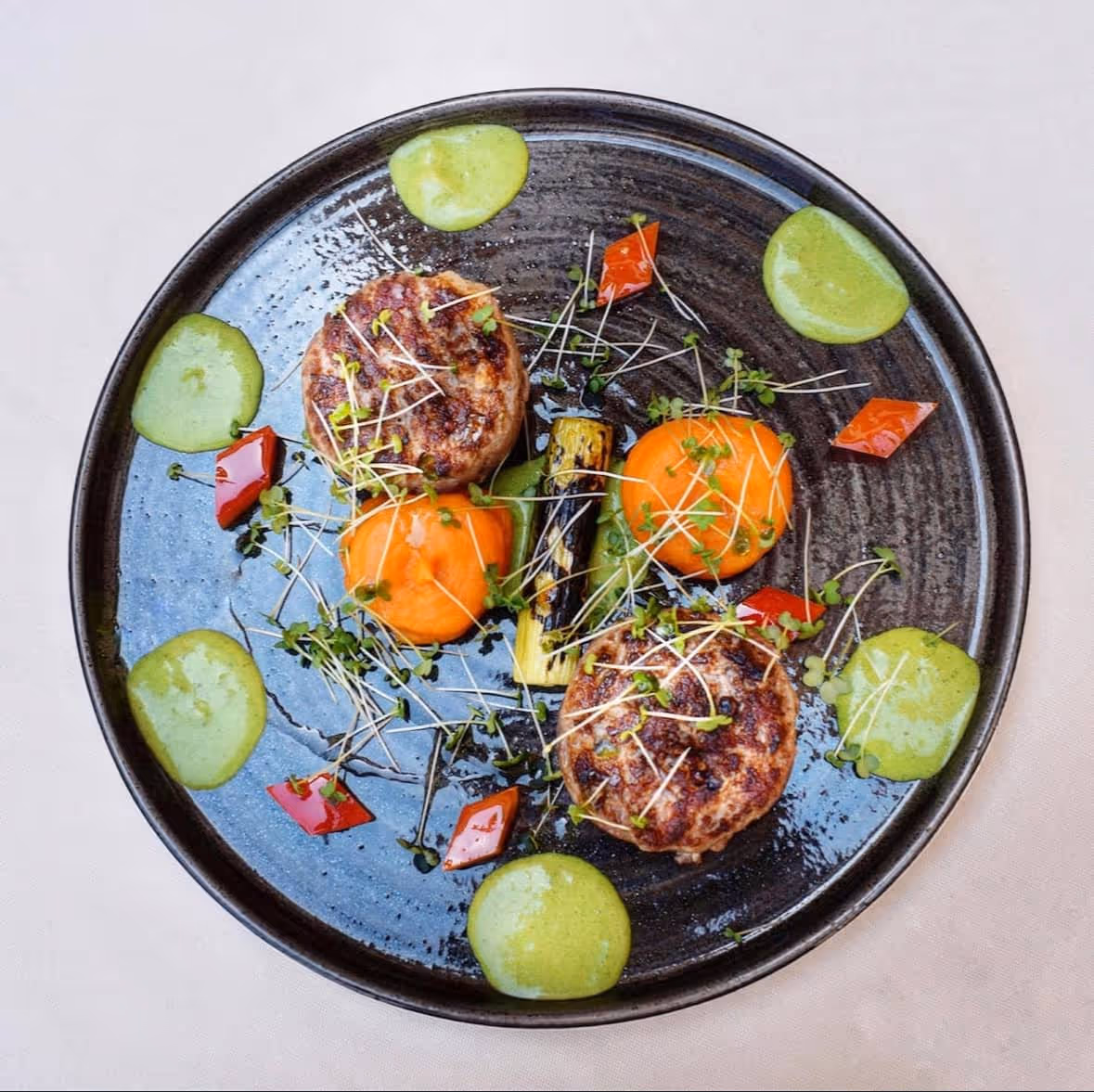 Grilled patties garnished with sprouts, served with dollops of green sauce, roasted vegetables, and red pepper pieces on a dark plate.