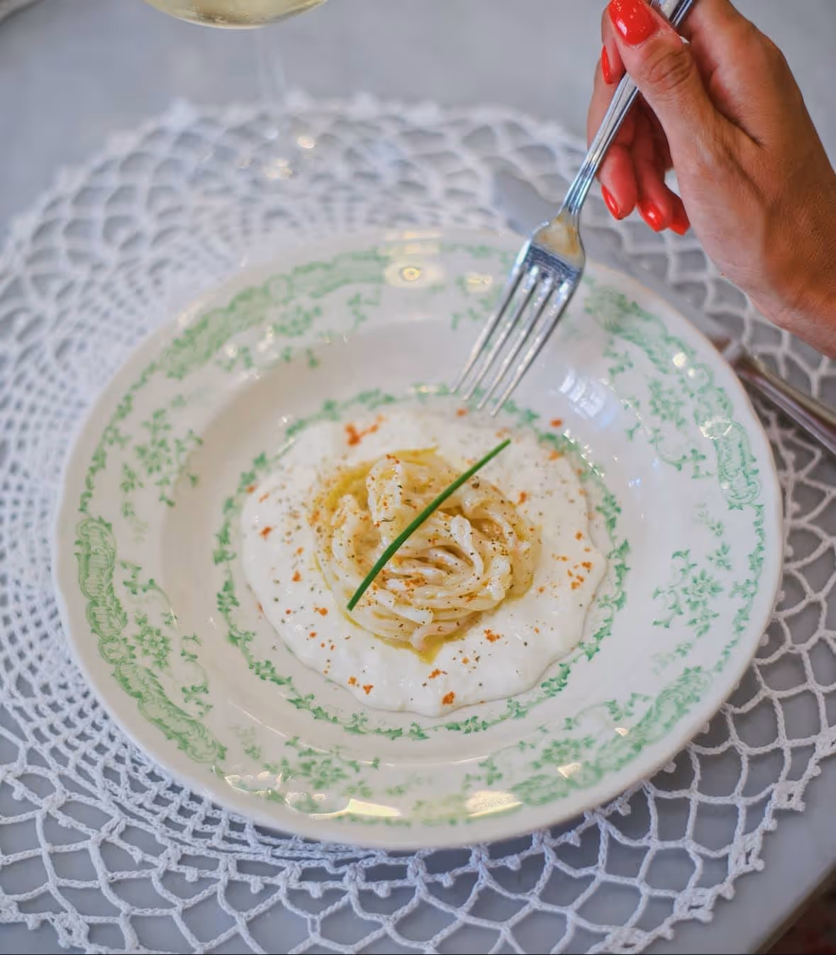 Plate of creamy pasta garnished with a chive, held by a hand with red-painted nails and a fork.