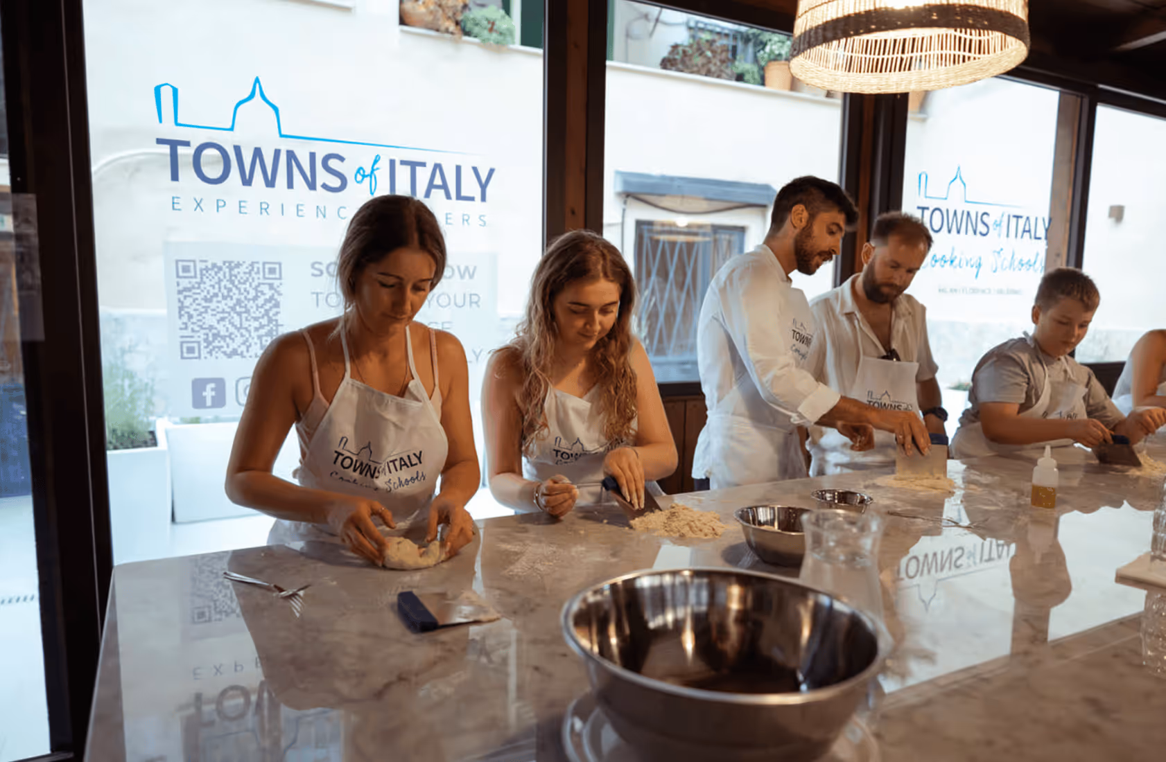 A group of people wearing Towns of Italy Cooking School aprons kneading and preparing dough on a marble counter inside a cooking class.