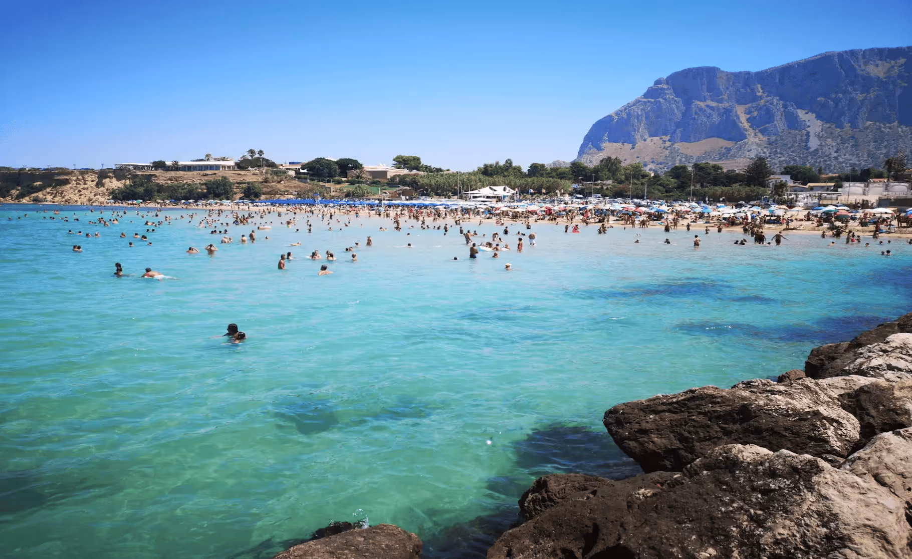 Crowded beach with people swimming in clear turquoise water and rocky foreground under a bright blue sky.