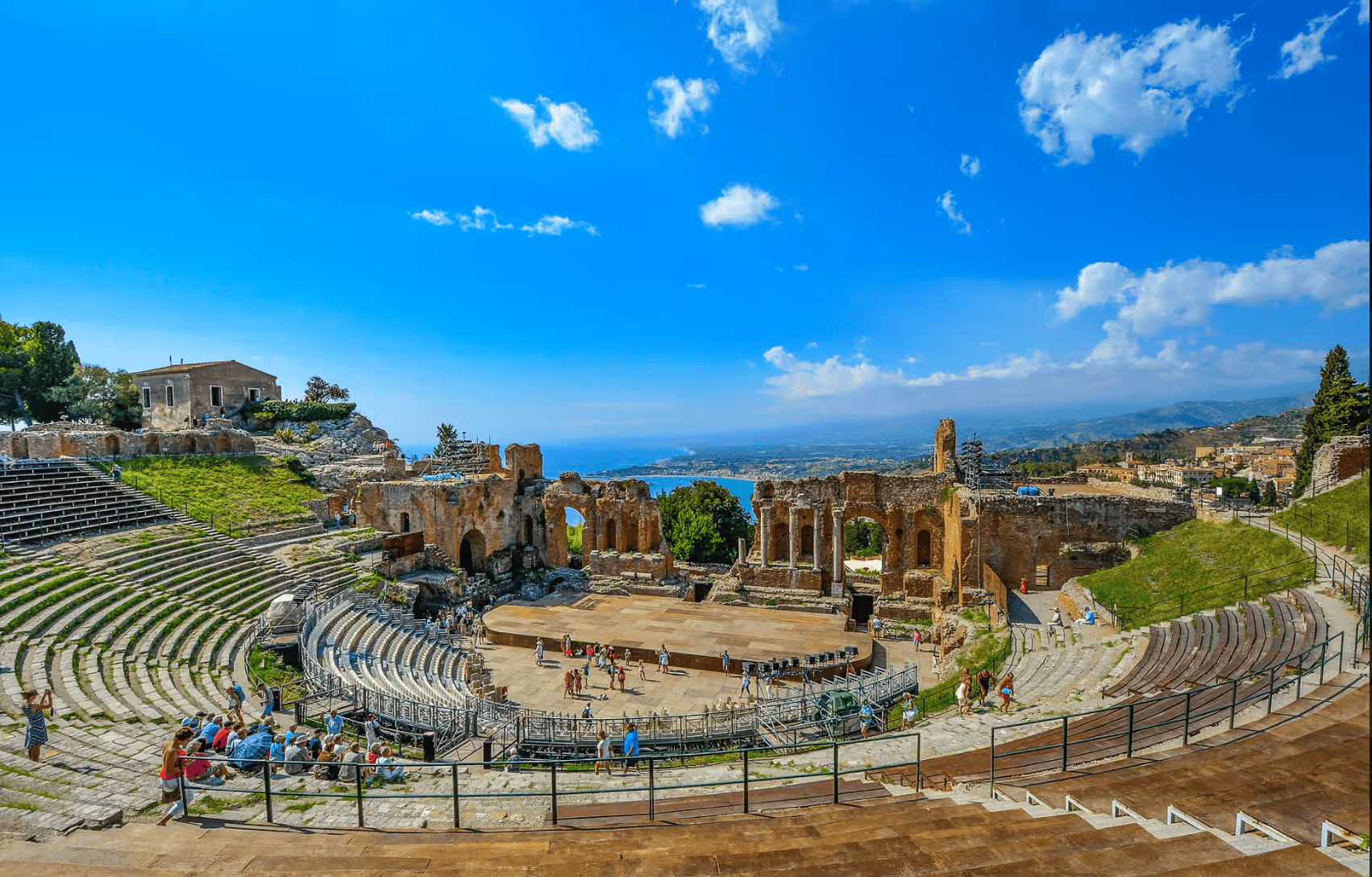 Ancient open-air amphitheater with stone seating and ruins under a bright blue sky, overlooking a coastal landscape.