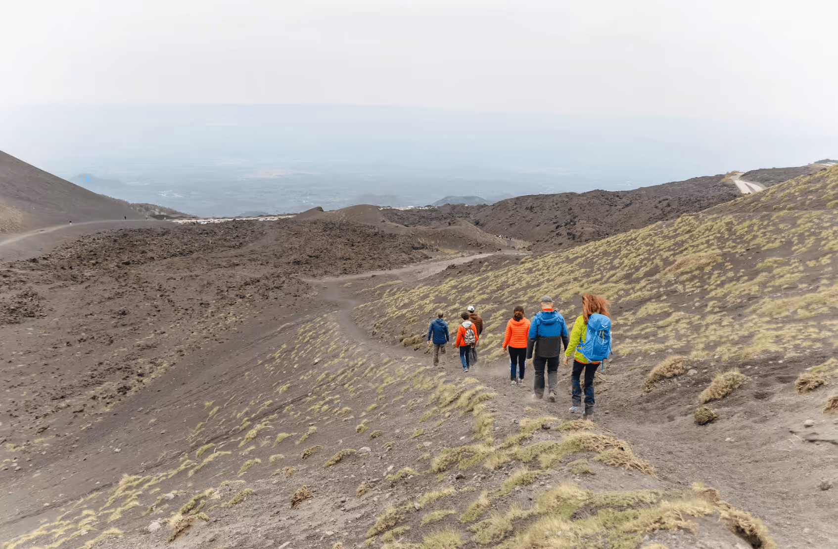 Group of six hikers walking down a barren, rocky mountain trail with sparse vegetation under an overcast sky.