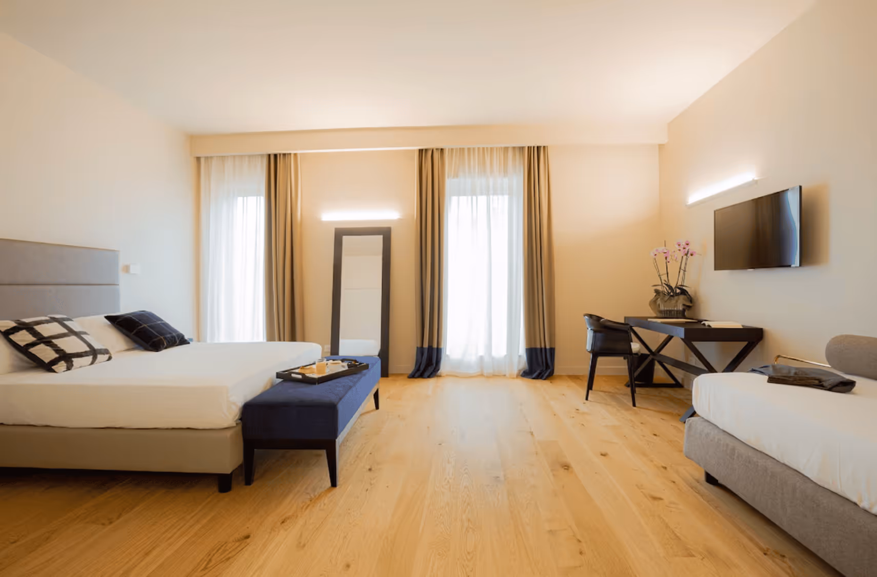 Bright modern hotel room with two beds, a navy bench, a wall-mounted TV, a desk with a chair, and large windows with curtains.