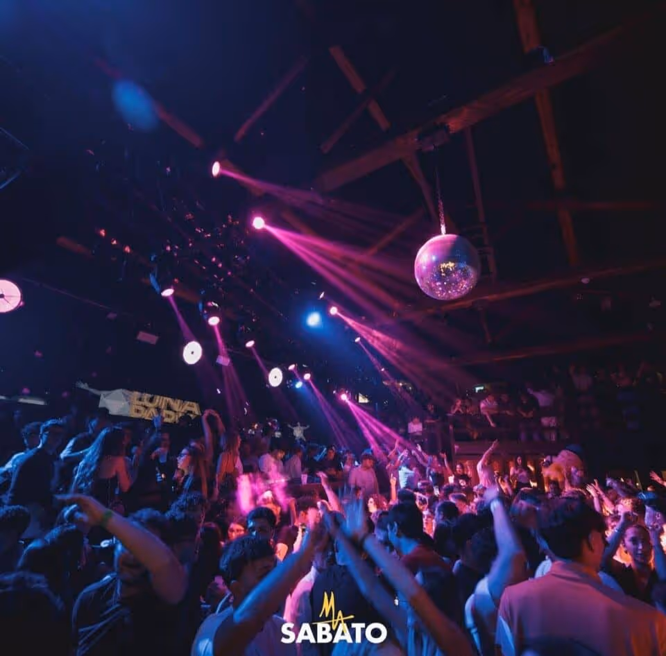 Crowded nightclub dance floor with people dancing under purple and blue spotlights and a hanging disco ball.
