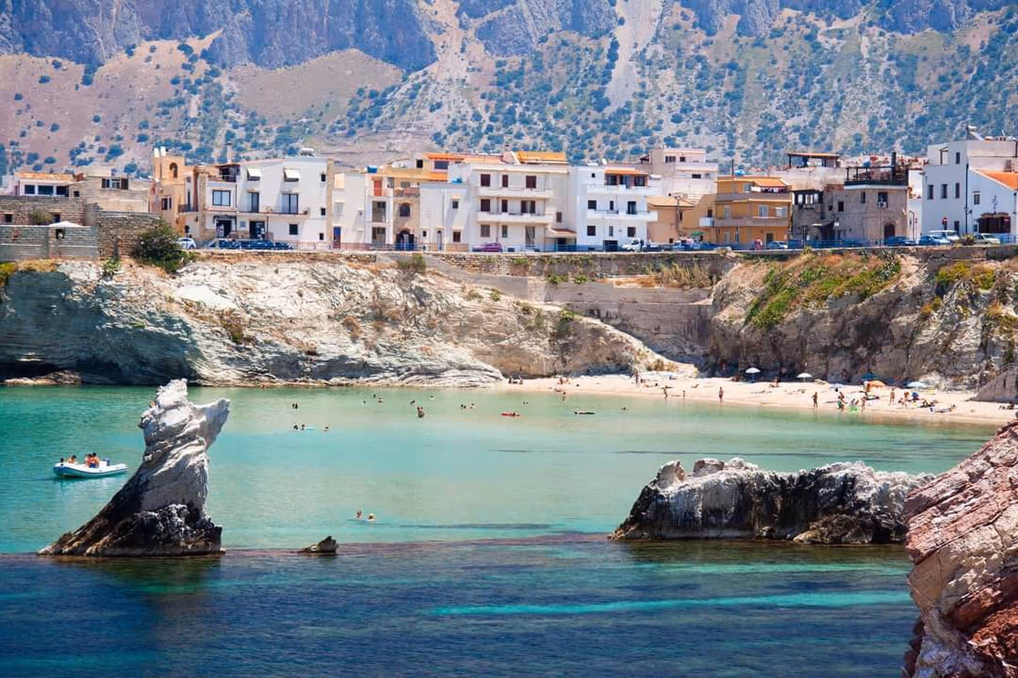 Coastal town with cliffs, turquoise water, small beach with people swimming and sunbathing, and buildings along the shoreline.