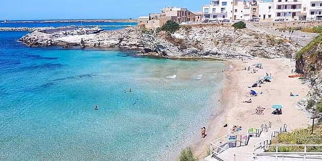 Clear turquoise sea beside a sandy beach with a few people, rocky cliffs, and buildings in the background.