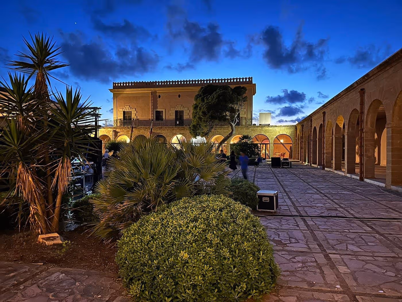 Stone courtyard with manicured bushes and palm trees in front of a historic building illuminated at dusk under a deep blue sky with scattered clouds.