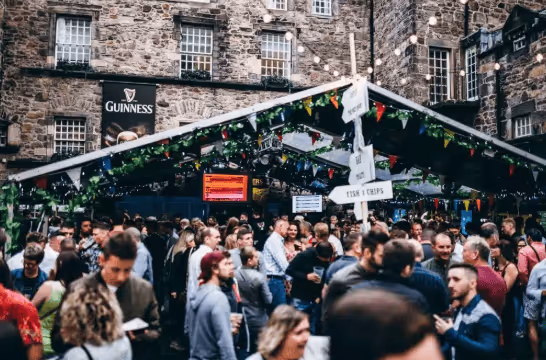 Crowd of people gathered under a tent with festive bunting and signs for food and drink outside a stone building with a Guinness banner.