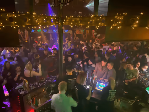 Crowded nightclub scene with DJ booth, colorful lights, and people dancing and socializing.