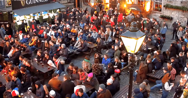 Crowd of people sitting and standing closely together at outdoor tables under a streetlamp near a Guinness stand.
