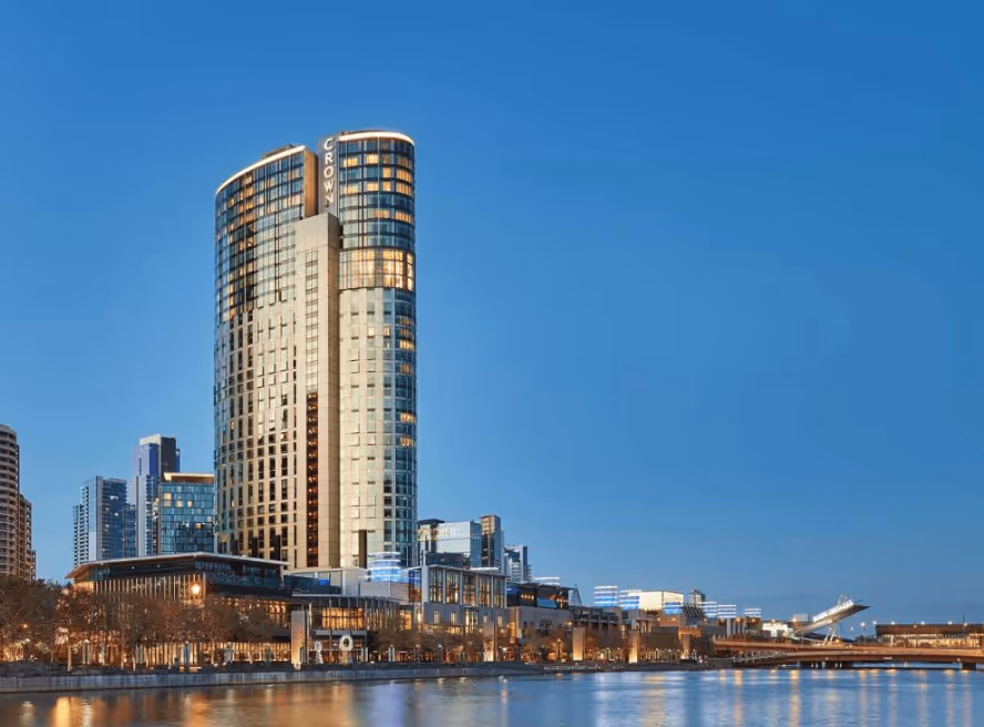 Tall modern Crown hotel building and cityscape along a waterfront at dusk with clear blue sky.